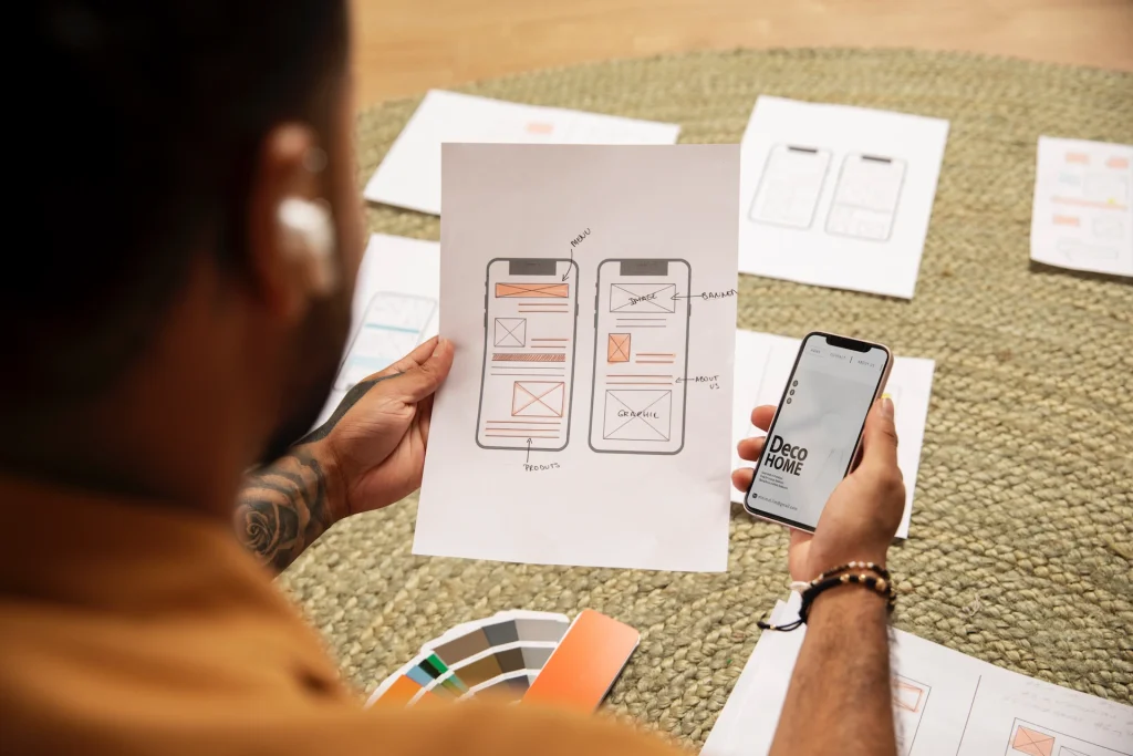 Designer comparing a printed wireframe sketch of a mobile app interface with its digital version on a smartphone